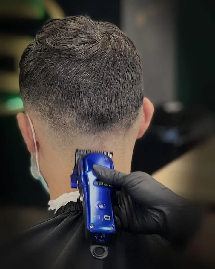 A barber in black gloves fading the back of a client's neck with a blue clipper
